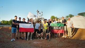 Grupowe zdjęcie zespołu AGH Space Systems pozuje do wspólnego zdjęcia podczas zawodów Anatolian Rover Challenge 2026. Uczestnicy trzymają flagę AGH, oraz flagę Polski. Pośrodku kilka osób z grupy podnosi nad głowami łazika.