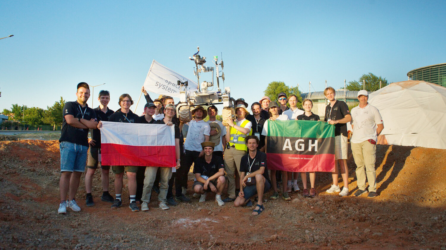 A group photo of the AGH Space Systems team during the Anatolian Rover Challenge 2026. The participants are holding an AGH University flag and a Polish flag. In the centre, several members of the group are lifting the rover above their heads.