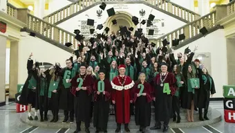 Grupowe zdjęcie wykonane w holu reprezentacyjnym budynku głównego AGH. Na schodach absolwenci i absolwentki w strojach akademickich wyrzucają w górę birety. W pierwszym rzędzie przedstawiciele władz rektorskich i dziekańskich trzymają w dłoniach cyfry 2024.