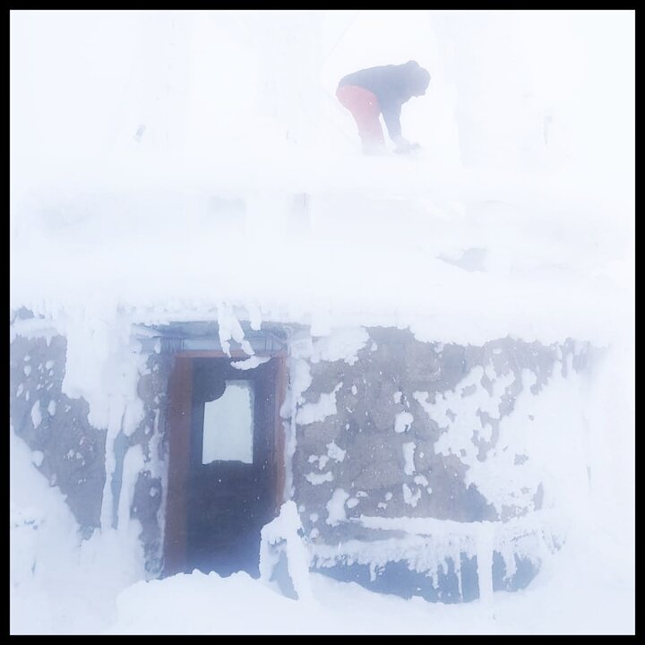 Wejście do Wysokogórskiego Obserwatorium IMiGW-PIB na Kasprowym Wierchu. Budynek przysypany jest grubą warstwą śniegu. Na dachu budynku stoi mężczyzna w schylonej pozycji, który zrzuca śnieg z dachu.