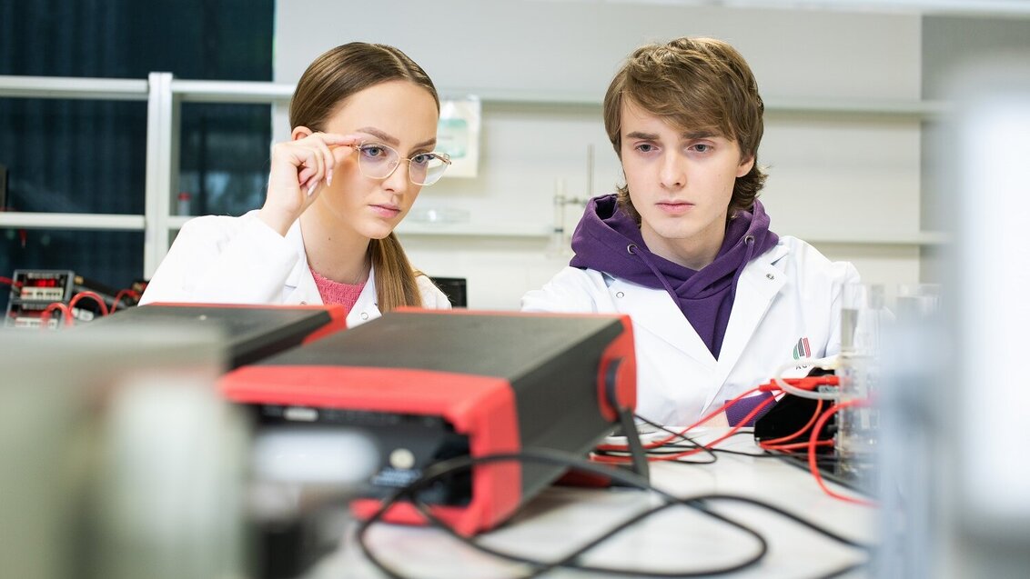 Na zdjęciu młodzi ludzie (kobieta i mężczyzna) w fartuchach laboratoryjnych. Siedzą obok siebie przy blacie, na którym rozłożony jest sprzęt (widać kable). Zdjęcie wykonane w sali ćwiczeń lub w laboratorium. 