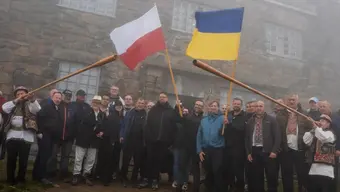 Group of people gathered outside a stone observatory building in misty mountain conditions, with the Polish and Ukrainian flags raised alongside traditional alpine horns held by participants in regional attire.