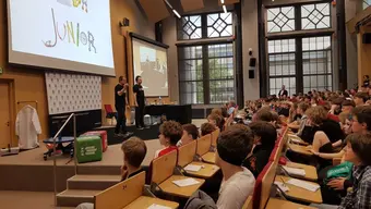 Sala wykładowa, w której siedzi licznie zgromadzona młodzież. Na scenie stoi dwóch naukowców, za nimi ekrany z napisami AGH Junior