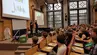 Sala wykładowa, w której siedzi licznie zgromadzona młodzież. Na scenie stoi dwóch naukowców, za nimi ekrany z napisami AGH Junior