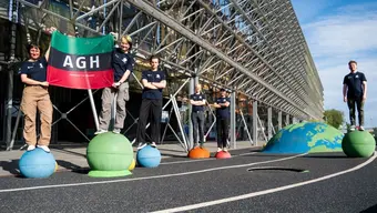 A group of students in matching T-shirts standing on colorful models of planets outside of an ESA facility. Two of them are holding the AGH University banner.