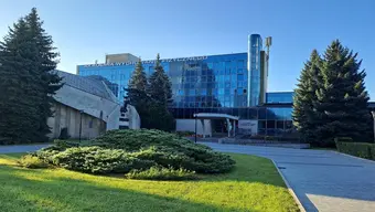 Widok na główny budynek Akademii Kultury Fizycznej w Krakowie, z przeszkloną fasadą, otoczony zielenią i drzewami, sfotografowany w słoneczny dzień.