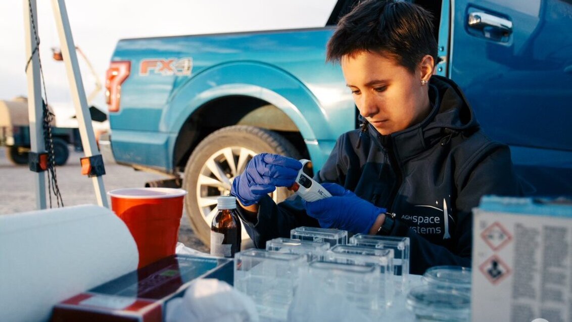 A female scientist prepares samples, behind her is a large blueish car.
