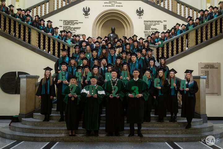 A commemorative photo of graduates and university authorities holding letters spelling "graduation" and numbers 2023, stading on the stairs in the main hall of the main AGH University building