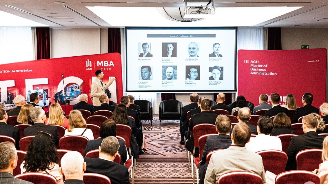 The participants of the MBA TECH inaugural meeting sit on chairs organised in a few rows. They are watching a presentation displayed on a large screen in the centre and listening to an elegantly dressed man delivering a speech. Next to the screen, there are temporary walls with MBA TECH logos.