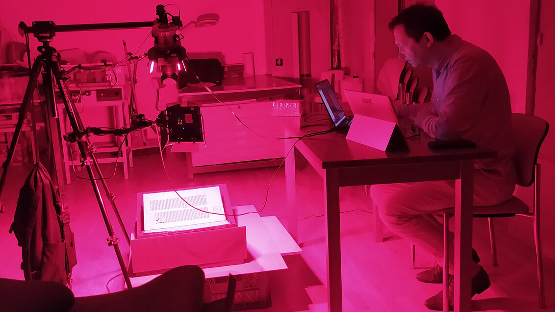 Image of the proces of determining lightfastness of a work. Dr Łojewski sitting at a desk on the right and the item being examined laying under bright pink light in the middle of the room.