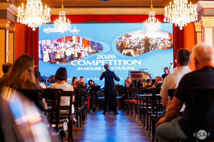 „Con Fuoco” wykonuje koncert podczas festiwalu w reprezentacyjnej sali koncertowej w Wiedniu. Na pierwszym planie dyrygent prowadzi zespół, a na widowni zgromadzona jest publiczność.