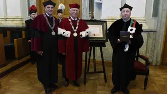 Trzy osoby w ceremonialnych togach akademickich pozują do wspólnego zdjęcia podczas uroczystości. W tle widoczny jest oprawiony dyplom na sztaludze.