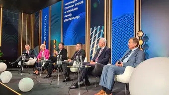 Prelegenci w trakcie dyskusji siedzą w fotelach ustawionych wzdłuż ściany. Wszyscy są elegancko ubrani. Przed nimi ustawiono niskie stoliczki, a na nich butelki z wodą. 