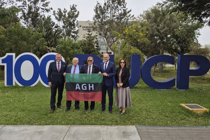 A group of university representatives posing with an AGH University flag in front of a large PUCP sign on a university campus.