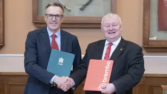 Na zdjęciu dwóch mężczyzn, którzy stoją obok siebie, trzymają w uścisku prawe dłonie, w lewych trzymają teczki z podpisanymi dokumentami.