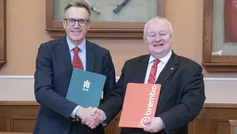 Image of two smiling men shaking hands, one holding a green AGH University folder, the latter a red one with the word "Brembo".