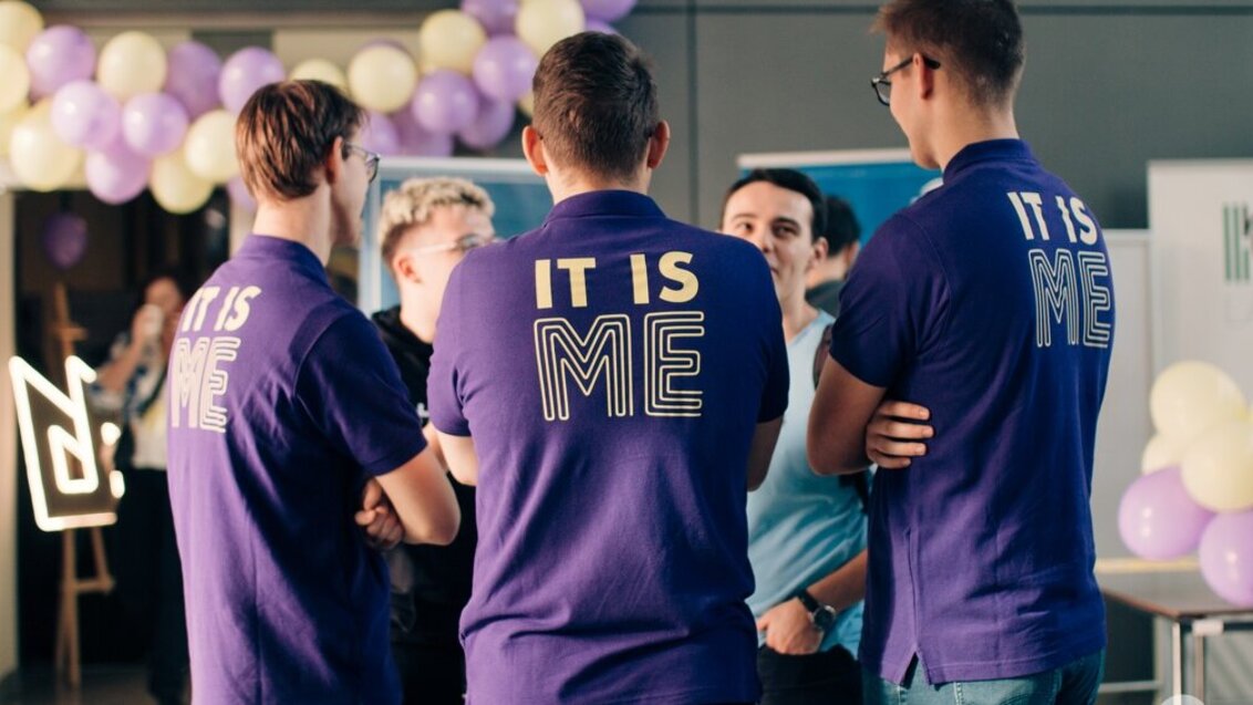 Zdjęcie z festiwalu IT is ME. Pięć osób stojących w kręgu rozmawia ze sobą. Trzy z nich stojące tyłem do obiektywu mają na sobie fioletowe koszulki z napisem „IT is ME”. W tle fioletowe i białe balony oraz plakaty.