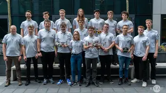 18 osób stojących w dwóch rzędach na tle nowoczesnego, przeszklonego budynku. Wszyscy mają ubrane jednakowe, jasnoszare koszulki z nadrukiem. Kilka osób trzyma w dłoniach drony lub elementy konstrukcyjne. W tle widać bezlistne drzewa.