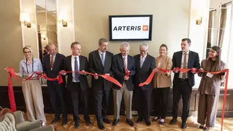 Uroczyste przecięcie wstęgi: dziewięć osób (kobiety i mężczyźni) stoi obok siebie trzymając w dłonicha czerwona wstęgę. Mężczyzna stojący pośrodku przecina wstęgę nożyczkami.