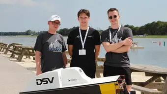 Na zdjęciu trzech młodych mężczyzn ubranych w t-shirty i shorty stoi na nabrzeżu wodnym. Przed nimi, na prowizorycznym podwyższeniu, autonomiczna łódź. R.O.B.U.R. Zdjęcie wykonane w słoneczny dzień.