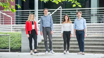 Four students, two women and two men, standing outdoors, in front of a flight of stairs.