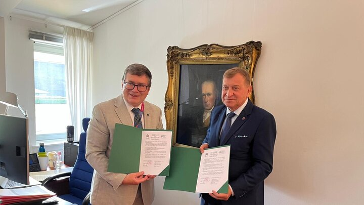 Two elegant men posing in front of an old, dark painting of a man in a decorative gold frame. They are holding open green folders with official documents.