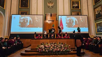 Na zdjęciu wnętrze auli AGH podczas uroczystości barbórkowych. na ekranach okolicznoścowy slajd, w stallach władze rektorskie i dziekańskie, przed stallami okazały bukiet kwiatów..