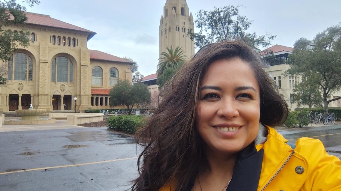 A smiling woman in a yellow rain jacket standing in the foreground of a Stanford campus courtyard.