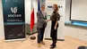 An image of a woman and a man shaking hands in front of the Polish and EU flags and two banners, one of the SOCCER project and the other of the NCBR.