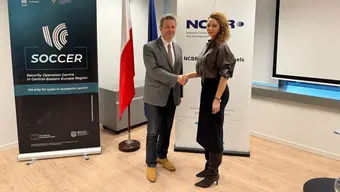 Dwie osoby podają sobie prawe dłonie w uścisku. Za ich plecami banery NCBR oraz projektu SOCCER. Pomiędzy banerami flaga Polski.