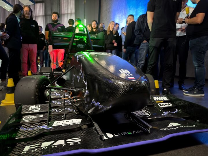 The front of the racing car, adorned with stickers including the AGH University logo, surrounded by spectators gathered in an industrial hall.