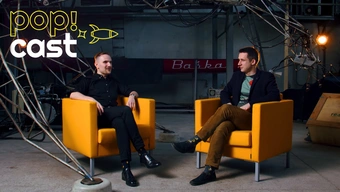 Zdjęcie przedstawia dwóch młodych mężczyzn rozmawiajcych ze sobą, siedzących w pomarańczowych fotelach wewnątrz hali. W pomieszczniu scenografię stanowi robotyczna rzeźba.