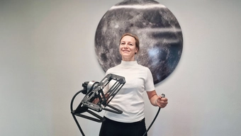 Kobieta w średnim wieku o blond włosach ubrana w białą bluzkę stoi na tle fotografii Księżyca trzymając w rękach urządzenie badawcze