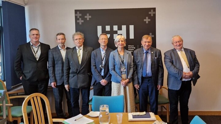 A group of professionally dressed individuals in a meeting room in front of a wall sign reading "HII." The setting includes a table with drinks and papers.