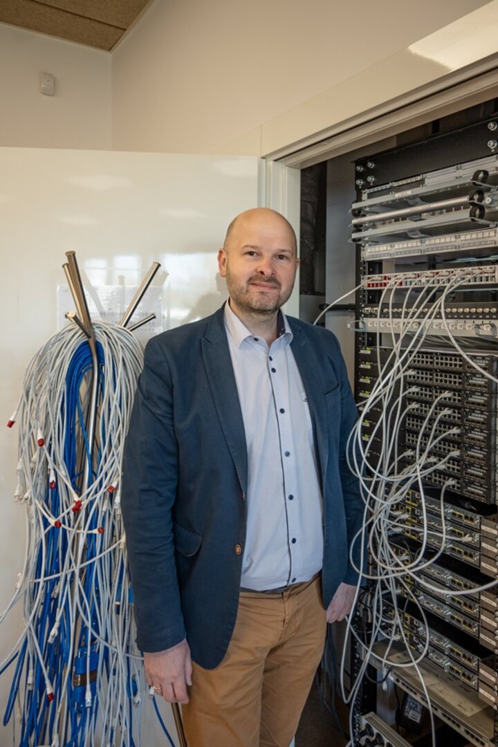 The photo shows AGH UST Professor Marcin Niemiec in a computer laboratory, posing against a set of servers and cables.
