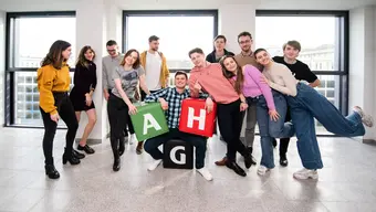 Na zdjęciu grupa młodych ludzi (studentek i studentów) stoją obok siebie w luźnych pozach, uśmiechają się. Pośrodku student siedzi na czarnym pufie z literą "G", trzyma na kolanach dwa pufy w kolorze zielonym i czerwonym z literkami "A" i "H". Zdjęcie wykonane w jednym z budynków AGH.
