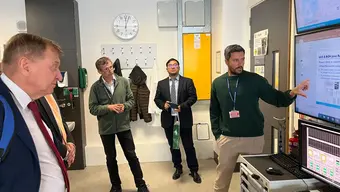 Wizyta w laboratorium. Cztery osoby stoją w pomieszczeniu z monitorami komputerowymi. Jedna z nich wskazuje na duży ekran z wyświetlanymi informacjami, pozostali obserwują.