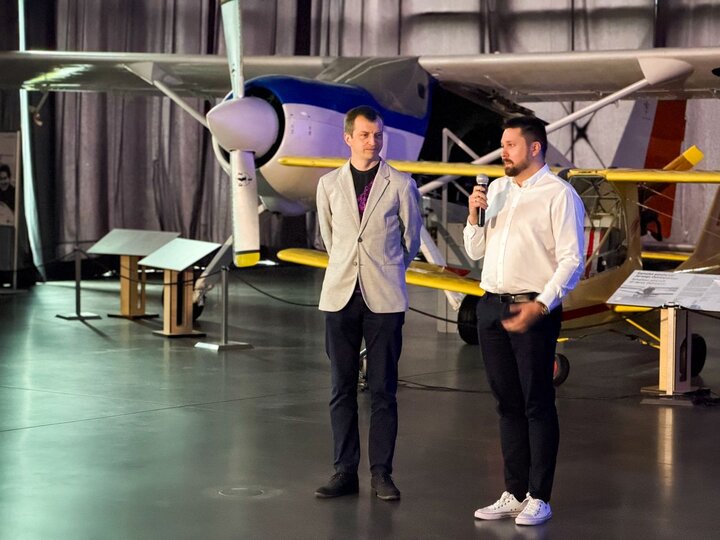 Two smartly dressed men, one of whom is speaking into a microphone. In the background, there is an aeroplane that forms part of the museum’s exhibition.