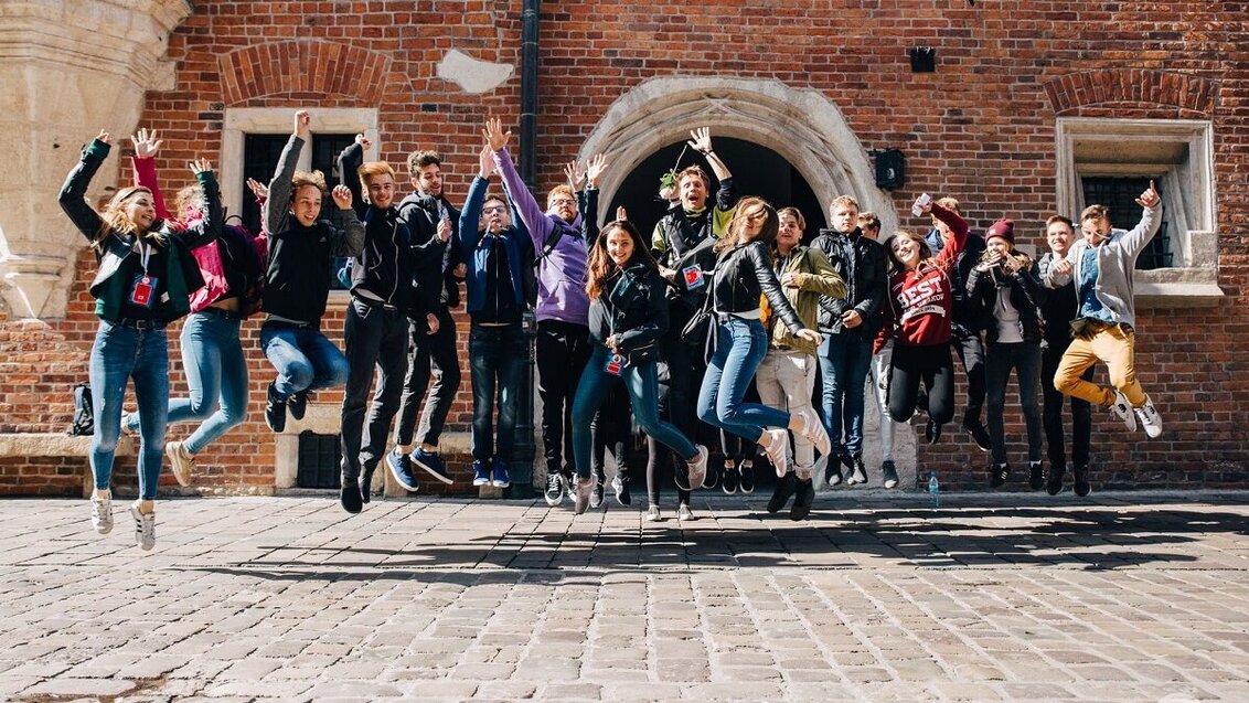 Na zdjęciu grupa kilkunastu młodych ludzi. Wszyscy podskakują , unosząc w górę ręce w geście radości. Zdjęcie wykonane na ulicznym placu, za plecami młodych ludzi widać ceglaną ścianę budynku.