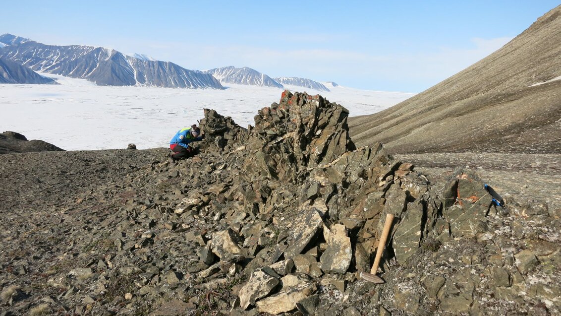 A woman examines rocks in the field, behind her is a layer of clouds, above which mountain peaks are visible.