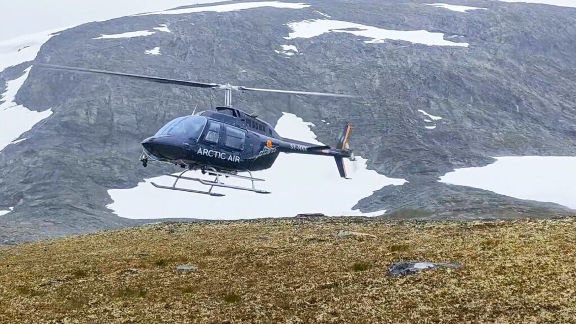 Helikopter z napisem ARCTIC AIR tuż nad powierzchnią ziemi. W tle widać góry z miejscami pokryte śniegiem.