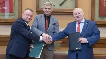 Sygnatariusze umowy (trzech mężczyzn w formalnych strojach) stoją obok siebie w sali konferencyjnej. Dwie osoby po bokach ściskają sobie dłonie, trzymając jednocześnie teczki z dokumentami. Osoba w środku stoi pomiędzy nimi, wspierając uścisk dłoni.