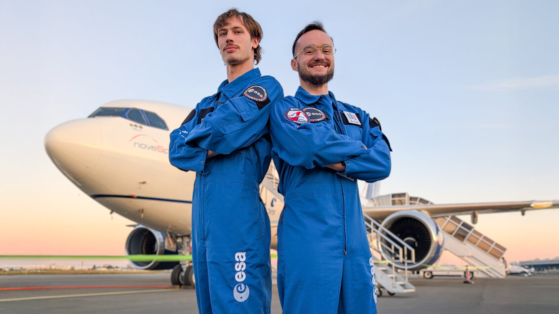 Two people in blue ESA suits standing in front of a large aircraft on the airport tarmac.