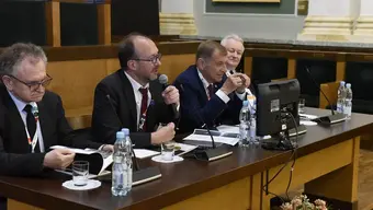 Aula AGH. Widoczne prezydium, przy którym siedzi czterech mężczyzn ubranych w garnitury. Jeden z nich trzyma w ręku mikrofon, inny - broszurę. Na stole: plastikowe butelki z wodą, szklanki, kartki, mikrofony, monitor komputera.