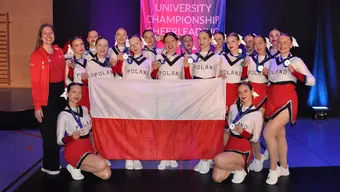 A group of cheerleaders in white and red uniforms posing for a photograph holding a Polish flag with their coach standing to the left.