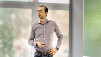 Na zdjęciu dr Jendrej stojący w sali wykładowej na tle dużych okien i tablicy z zapisanymi wzorami matematycznymi. W dłoni trzyma pilota do prezentacji.