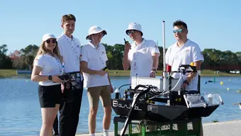 Zdjęcie członków zespołu (pięć osób) oraz łodzi Barka na tle akwenu wodnego. Młodzi ludzie ubrani są w identyczne, białe koszulki polo z logotypem AGH Solar Boat. Zdjęcie wykonane w słoneczny dzień.