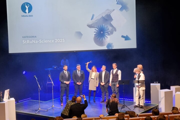 Zdjęcie z gali konkursu. Na scenie stoi kilka osób. Jedna z nich trzyma w podniesionej dłoni pamiątkowa statuetkę.