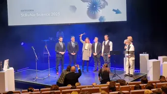 Zdjęcie z gali konkursu. Na scenie stoi kilka osób. Jedna z nich trzyma w podniesionej dłoni pamiątkowa statuetkę.