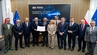 Representatives of partner institutions pose for a group photograph during the signing of a cooperation agreement, holding the signed document in front of a display screen and national and institutional flags.
